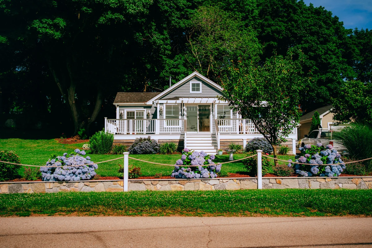Small white rural home framed by flowers, a picket fence, and leafy trees.
